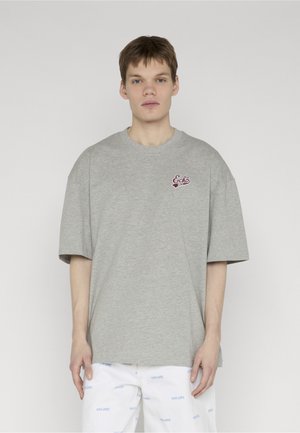 Mladý muž v voľnom sivom tričku s logom "Ecko" na hrudi a bielych nohaviciach s modrým vzorom textu "ecko unltd.".