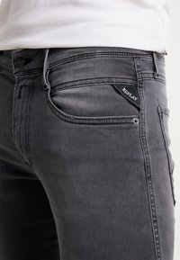 Vaqueros de mezclilla gris con un ajuste entallado, que cuentan con una etiqueta en el bolsillo trasero, trabillas para cinturón y cierre de botón. Detalles sutiles de textura y costura visibles.