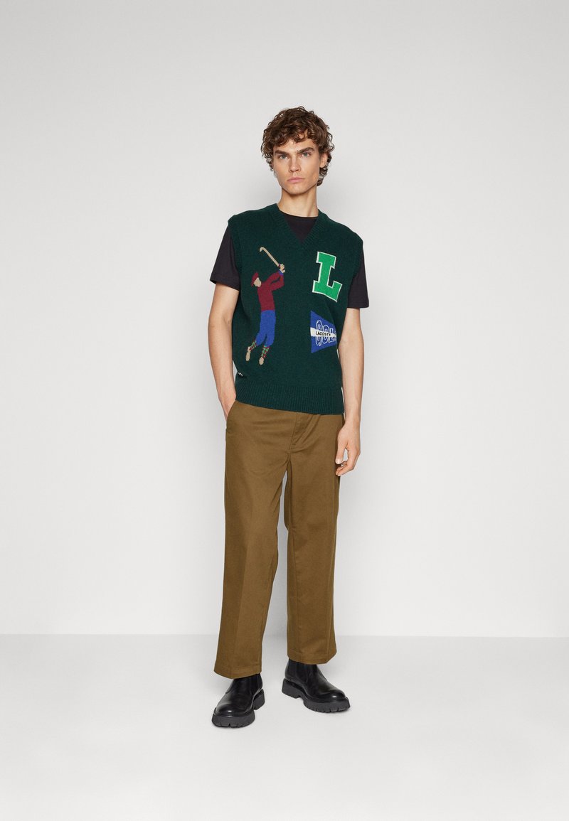 Gilet verde lavorato a maglia con grafica di un golfista colorato e lettera "L", indossato sopra una maglietta nera; abbinato a pantaloni marroni e scarpe nere.