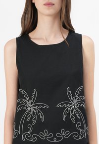 Femme portant un haut noir sans manches avec des motifs brodés blancs de palmier et de vagues près de l'ourlet, recadrée du menton à la taille.