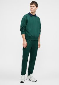 Felpa verde con colletto blu navy, abbinata a pantaloni da jogging verdi con righe laterali, indossata con scarpe da ginnastica grigie e bianche.