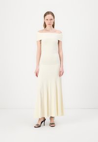 Robe légère crème décolletée avec un corsage ajusté et une jupe plissée fluide. Des sandales à talons noires avec des lanières complètent la tenue.