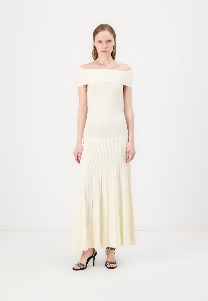 Robe légère crème décolletée avec un corsage ajusté et une jupe plissée fluide. Des sandales à talons noires avec des lanières complètent la tenue.