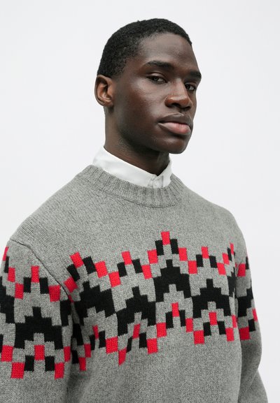 Νεαρός άνδρας ντυμένος με γκρι πουλόβερ με κόκκινα και μαύρα γεωμετρικά σχέδια, από πάνω από ένα κουμπωτό λευκό πουκάμισο, ποζάροντας μπροστά από ένα απλό λευκό φόντο.