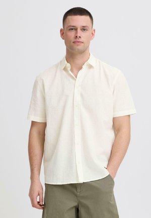 Ung mand iført en hvid, kortærmet tekstureret skjorte med knapper og olivengrønne bukser, stående med den ene hånd i lommen.