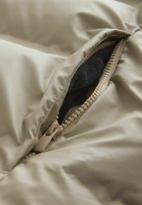 Jachetă bej căptușită dintr-un material cu fermoar vizibil; interiorul căptușit cu material negru. Textură moale cu elemente de design matlasate.