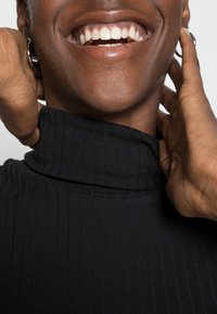 Haut à col roulé noir côtelé avec un col haut, un design ajusté et une texture lisse, entouré de mains et d'une bouche souriante.