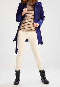 Navy blue waterproof trench coat with buttons over a tan striped shirt, paired with cream pants and black ankle boots with buckles.