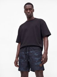 Mann trägt ein schwarzes oversized T-Shirt und dunkelblaue Camouflage-Shorts mit Kordelzug und einem braunen Logo-Patch auf weißem Hintergrund.