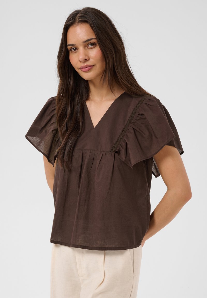 Jeune femme aux longs cheveux foncés portant un chemisier marron ample à col en V avec des manches courtes volantées et un pantalon couleur crème, debout devant un fond uni.