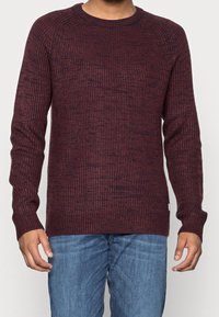 Bordeauxfärgad stickad tröja med en texturerad, ribbad design. Har långa ärmar och en klassisk rundhalsad krage. Bärs med slim-fit blå jeans.