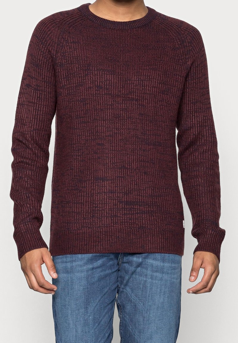 Bordeauxfärgad stickad tröja med en texturerad, ribbad design. Har långa ärmar och en klassisk rundhalsad krage. Bärs med slim-fit blå jeans.