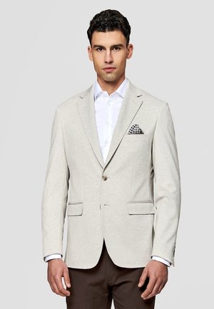 Mężczyzna w beżowej marynarce z wzorzystą poszetką, białą koszulą i brązowymi spodniami, stojący na jednolitym jasnoszarym tle.