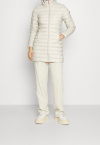 Lysfarvet pufferjakke med vandret quiltet design, høj krave og lynlås foran. Båret med beige bukser og chunky hvide sneakers.