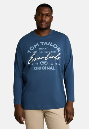 Mężczyzna w niebieskiej koszuli z długim rękawem z białym napisem "Tom Tailor Essentials Original" i beżowych spodniach, stojący na tle jednolitego jasnego tła.