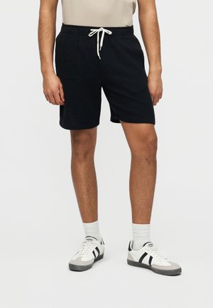 Άνδρας που φοράει μαύρα σορτς με κορδόνι, λευκές κάλτσες και λευκά αθλητικά παπούτσια με μαύρες ρίγες σε απλό φόντο.