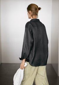 Camicia nera di lino con maniche lunghe e vestibilità rilassata, abbinata a pantaloni verde chiaro e con una borsa bianca strutturata.