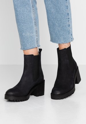 Bottines noires à talon épais portées avec un jean court bleu clair aux ourlets effilochés sur un fond clair uni.