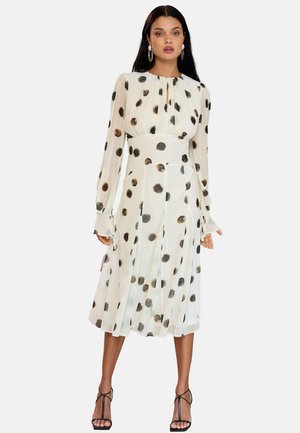 Nő, aki fehér midi ruhát visel fekete és barna pöttyökkel, hosszú ujjú, deréknál húzott, fekete pántos magas sarkú cipővel.