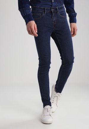 Ciemnoniebieskie skinny jeans z widocznym przeszyciem i pięcioma kieszeniami, noszone z białymi sneakersami. Materiał to gładki denim, dopasowany krój.