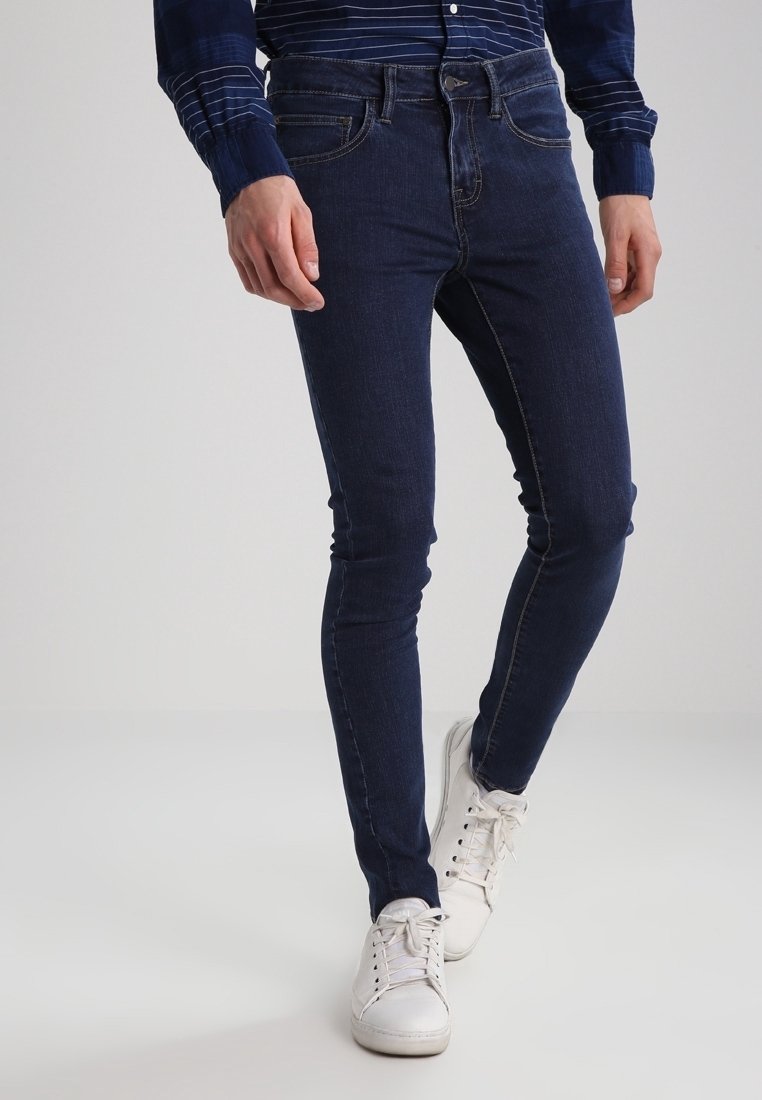 Tmavě modré skinny džíny s viditelným švem a pěti kapsami, nošené s bílými teniskami. Textura je hladký denim, těsný střih.