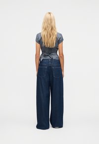 Femme blonde vue de dos, portant un chemisier gris à manches courtes noué et un jean bleu foncé à jambes larges, sur un fond uni.