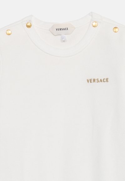 T-shirt blanc pour bébé avec des détails de boutons dorés sur les épaules et petit logo doré Versace sur la poitrine.