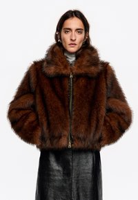 Brązowa kurtka z sztucznego futra z szerokim kołnierzem, zamkiem z przodu i miękką teksturą. Posiada krótki fason z obszernymi rękawami i czarnymi skórzanymi akcentami.