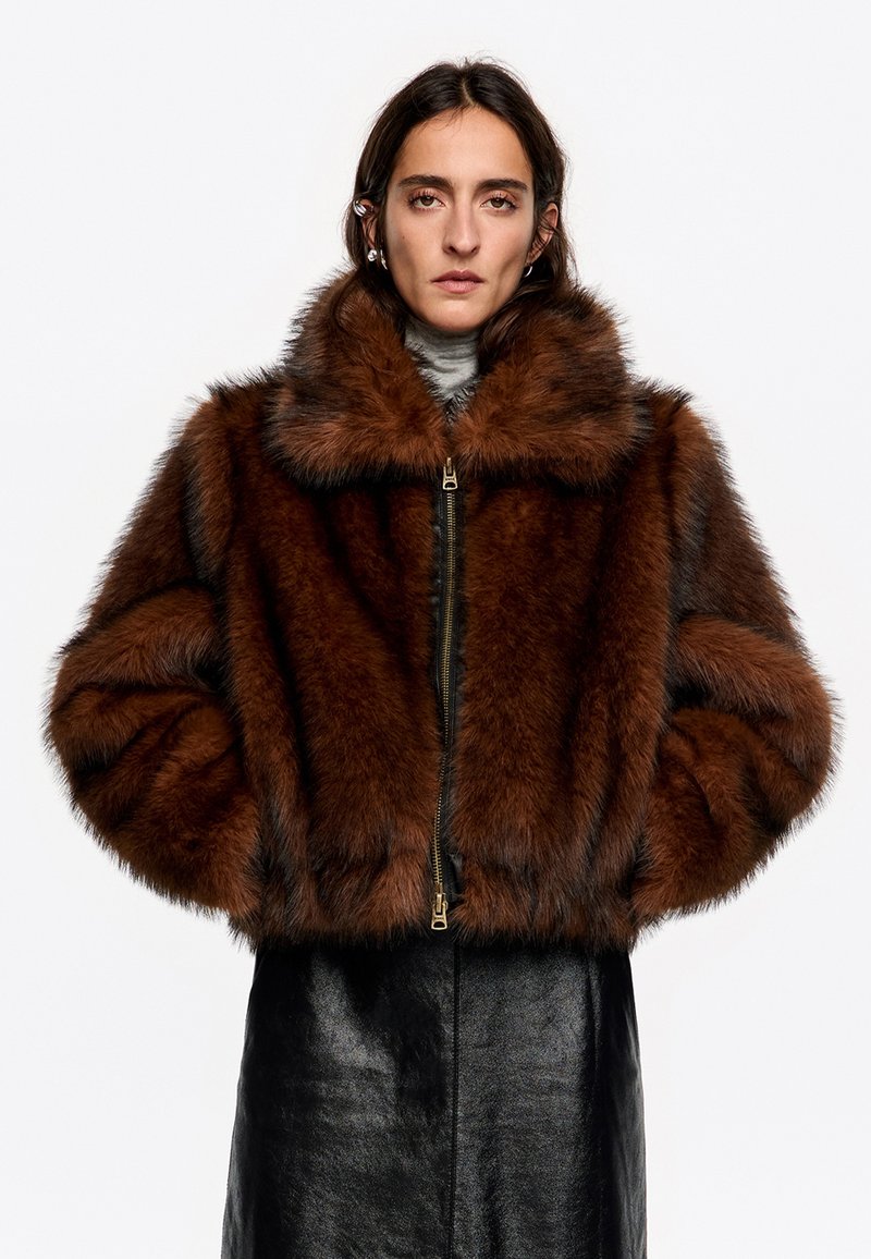Brązowa kurtka z sztucznego futra z szerokim kołnierzem, zamkiem z przodu i miękką teksturą. Posiada krótki fason z obszernymi rękawami i czarnymi skórzanymi akcentami.