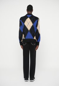 Man staat met zijn rug naar de camera, gekleed in een zwart, crème en blauwe ruitvormige trui, zwarte jeans en zwart-witte sneakers.