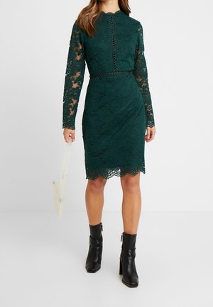 Frau trägt ein dunkelgrünes Spitzenkleid mit langen Ärmeln in knielanger Länge, schwarze Stiefeletten und hält eine kleine weiße Handtasche mit Perlen.