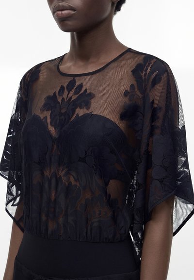 Personne à la peau foncée portant un haut en dentelle noire transparente avec des motifs floraux et un col rond, laissant apparaître un vêtement sans bretelles en dessous.
