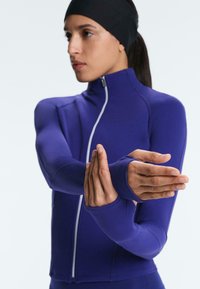 Femme portant une veste zippée violette ajustée et un bandeau noir, étirant ses bras à travers sa poitrine, se préparant à l'exercice.