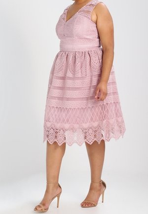 Donna con indosso un vestito rosa di pizzo senza maniche, lungo fino al ginocchio, con motivi intricati e sandali beige con tacco alto, in piedi davanti a uno sfondo semplice.