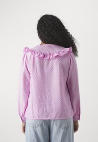 Blusa rosa chiaro con scollatura arricciata e maniche lunghe, tessuto testurizzato con un sottile motivo a quadretti, abbinata a jeans di denim.
