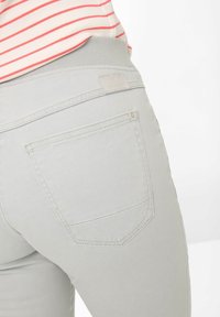 Lichtgrijze jeans met een taps toelopende pasvorm, achterzakdetail en elastische tailleband. Het materiaal lijkt zacht met minimale stikselaccenten.