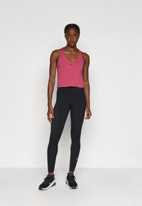 Ružový športový crop top s výstrihom do V a logom, spárovaný s čiernymi legínami s vysokým pásom a čiernymi športovými topánkami. Jednoduchý dizajn, priliehavý strih.