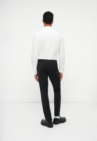 Weißes Langarmhemd, schwarze Slim-Fit-Hosen und schwarze Schuhe, von hinten vor einem schlichten weißen Hintergrund gezeigt.