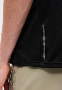 Detailní záběr paže osoby vedle její černé košile s vertikálním reflexním pruhem a béžových kalhot s rukou v kapse.