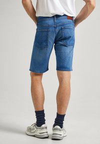 Pantalones cortos de mezclilla en azul medio con corte recto, cinco bolsillos y una etiqueta de cuero en la cinturilla. Combinados con zapatillas grises y calcetines tobilleros azul marino.