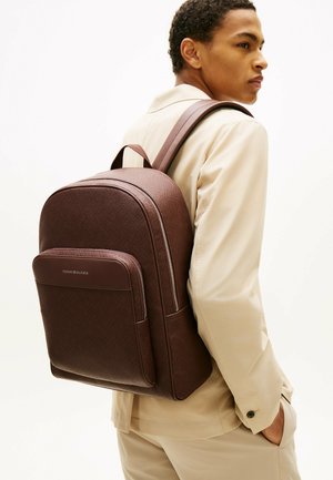 Jeune homme portant un costume beige et un sac à dos en cuir marron foncé texturé, avec une poche zippée à l'avant et le logo Tommy Hilfiger.