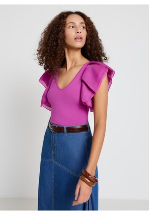 Donna con capelli ricci che indossa un top senza maniche magenta con spalle arricciate, gonna in denim blu, cintura marrone e braccialetti impilati.