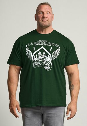 Homme aux cheveux courts et barbe, portant un t-shirt vert foncé avec un motif de moto blanc et le texte "L.A Custom Parts", debout devant un fond uni.