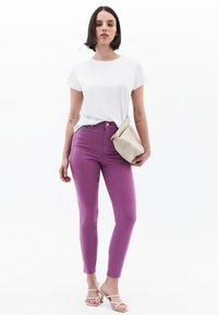 Jeans skinny morados, diseño ajustado, combinados con una camiseta blanca. Sosteniendo un bolso de mano de color gris claro. Sandalias sencillas completan el look.