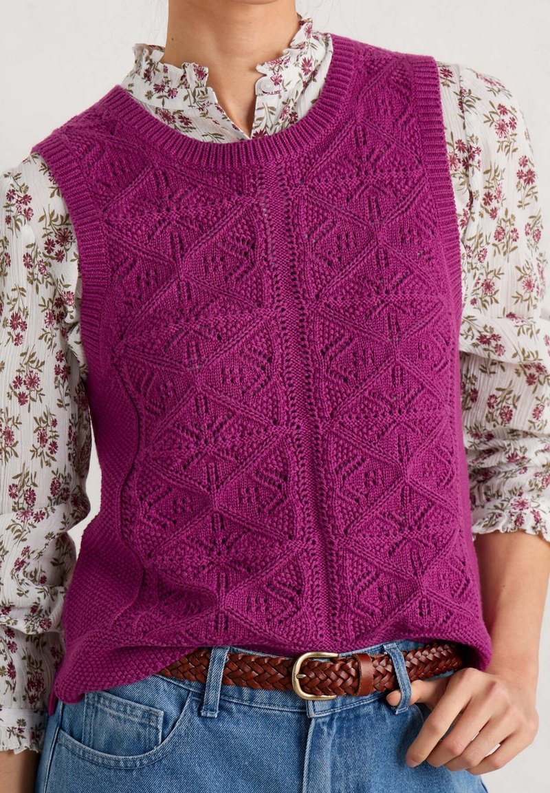 Femme portant un gilet sans manches tricoté violet sur un chemisier blanc à motif floral avec des manches bouffantes et un jean bleu avec une ceinture tressée marron.