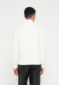 Mann in einem weißen, hochgeschlossenen Strickpullover und schwarzen Lederhosen, der mit dem Rücken zu einem schlichten weißen Hintergrund steht.