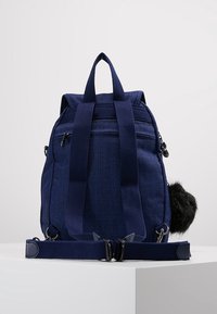 Sac à dos en tissu bleu marine avec des bretelles réglables, une poche zippée à l'avant et un porte-clés en fausse fourrure noire attaché sur le côté.