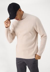 Beige Rippschal mit langen Ärmeln, kombiniert mit einer schwarzen Baseballkappe. Der Pullover hat einen lockeren Schnitt und eine strukturierte Oberfläche.