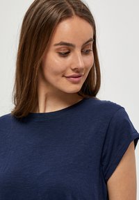 La camiseta azul marino presenta mangas cortas enrolladas, un cuello redondo y una tela suave y texturizada. Diseño casual adecuado para el uso diario.
