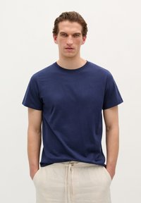 Marineblå t-shirt i blødt stof, rund halsudskæring, korte ærmer, diskret logo detalje på brystet, båret afslappet med lyse bukser.